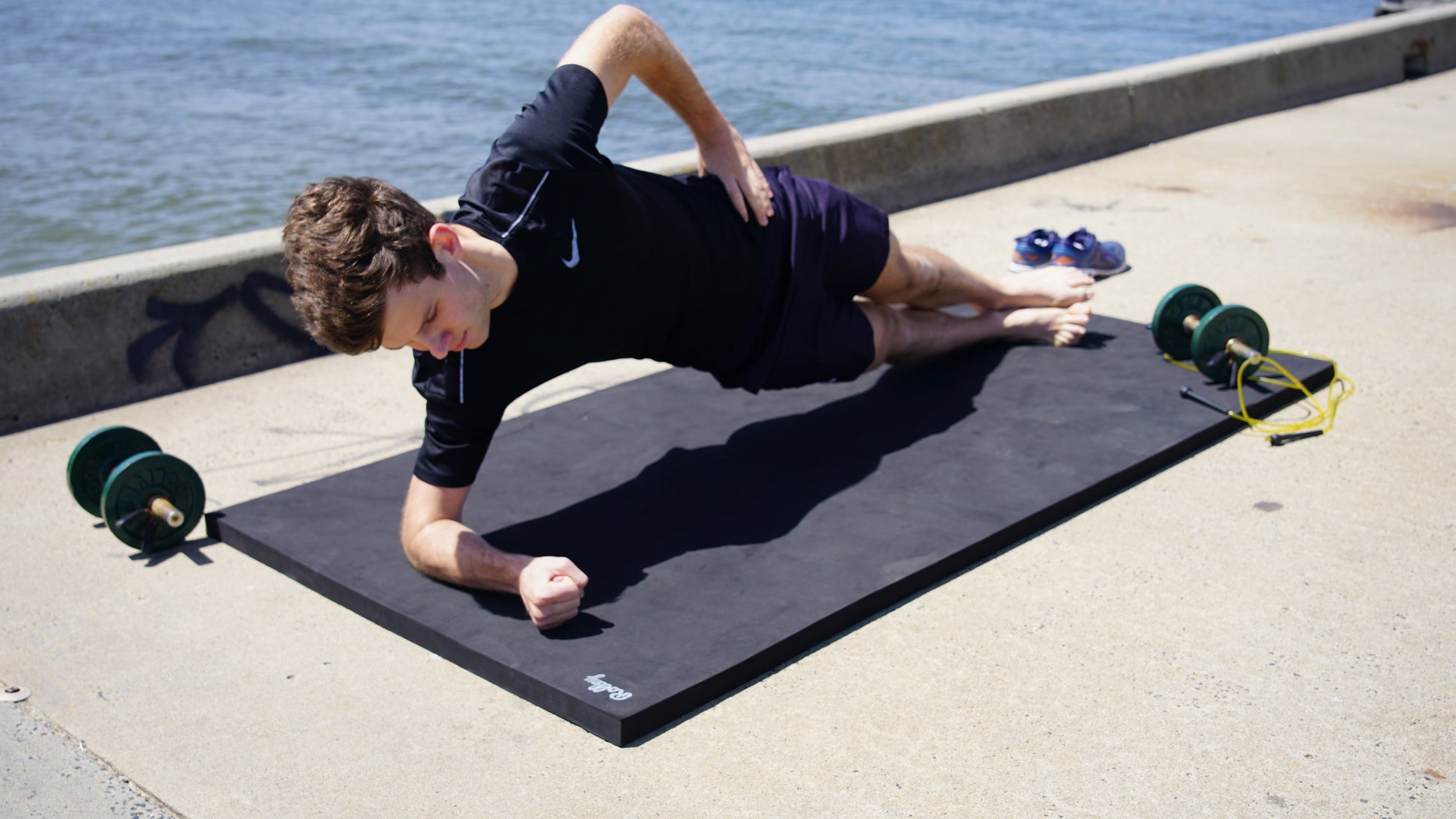 Person exercising on a Rolley Max mat beside dumbbells and a jump rope by the water